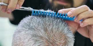 Ein Mann mit ergrautem Haar ist beim Friseur. Mit einem blauen Kamm und einer Haarschere schneidet die Friseurin die Haarspitzen des Herrn.