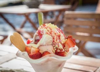 In einer Eisdiele, an einem herrlichen, sonnigen Tag steht ein Eisbecher mit Erdbeeren und Erdbeersauce auf dem Tisch. Es befindet sich in einem schönem Glas mit einem Plastiklöffel darin.