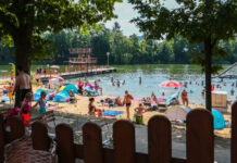 Ein Badesee mit einem Strandbad in Deutschland, in dem sich viele Gäste tummeln. Einige schwimmen im Wasser, andere liegen am Strand und sonnen sich.