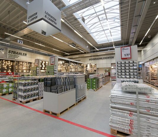 Blick in einen Baumarkt in Deutschland. Vorne stehen verschiedene Materialien, die man für Haus und Garten benötigt. Im Hintergrund erkennt man die Abteilung für erschiedene Arten von Lampen und Leuchtmitteln.