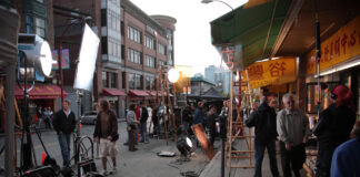 Auf einer Straße in der Stadt findet ein Filmdreh statt. Um den Film zu produzieren, stehen zahlreiche Lampen und Kameras bereit. Auf der Straße steht auch das Film-Team für den Dreh.