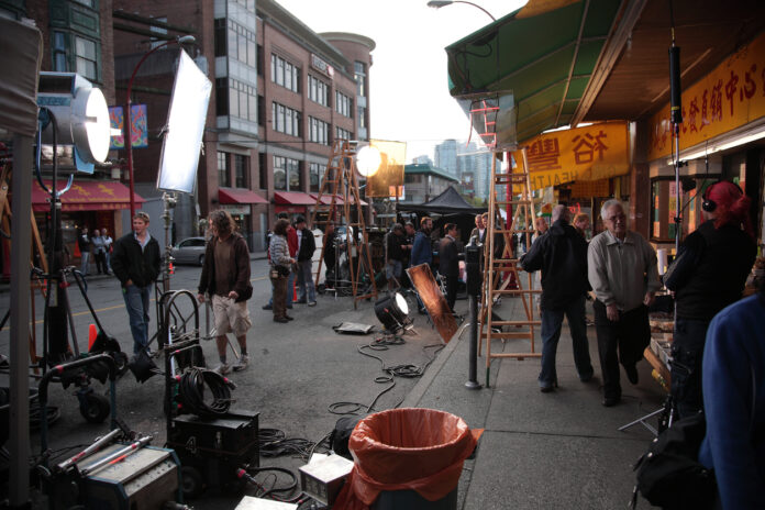 Auf einer Straße in der Stadt findet ein Filmdreh statt. Um den Film zu produzieren, stehen zahlreiche Lampen und Kameras bereit. Auf der Straße steht auch das Film-Team für den Dreh.