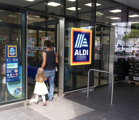 Aldi Eingang mit Familie. Ein Vater betritt mit seiner Tochter an der Hand gerade eine Aldi-FIliale. Vor der Filiale steht eine Holzpalette mit vielen Säcken Grillkohle.