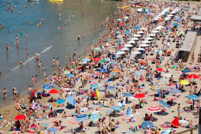 Ein stark gefüllter Strand, der mit vielen Badegästen und ihren Sonnenschirmen bestückt ist. Der Tag ist sommerlich und das Badewasser gut besucht.