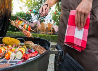 Ein Mann steht im Garten an einem Grill und hält mit einer Grillzange einen Spieß mit Gemüse und Fleisch hoch. Auf dem Grill liegen Gemüsespieße, Würstchen und Grillfleisch.