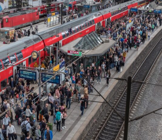 Ein extrem überfüllter Bahnhof von oben fotografiert. Die Menschenmenge tummelt sich auf dem Bahnsteig zwischen den Gleisen, um eine Regionalbahn zu erwischen. Ein roter Zug steht im Bahnhof, bereit zur Abfahrt.