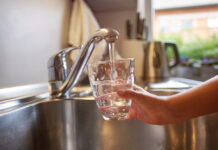 Eine Person steht vor dem Waschbecken und füllt ein Glas mit Wasser aus dem Wasserhahn. Dahinter ist eine beige Küche mit Messern und einem Wasserkocher abgebildet.