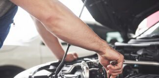 Ein Mann hält Werkzeug in den Motorraum eines Autos. In der Werkstatt steht im Hintergrund noch ein weiteres Fahrzeug in Weiß.