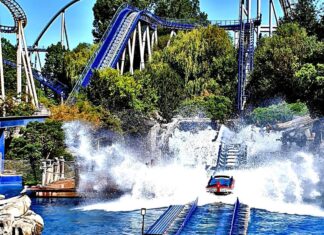 Mehrere Besucher sitzen in einer Art Boot auf einer Achterbahn mit dem Namen Poseidon im Europa-Park Rust. Die Schienen des Fahrgeschäfts befinden sich im Wasser und eine riesige Fontaine spritzt an der Seite in die Hlhe.