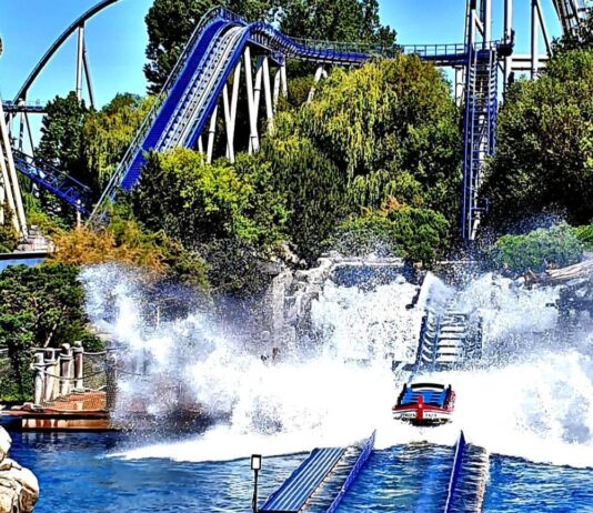 Mehrere Besucher sitzen in einer Art Boot auf einer Achterbahn mit dem Namen Poseidon im Europa-Park Rust. Die Schienen des Fahrgeschäfts befinden sich im Wasser und eine riesige Fontaine spritzt an der Seite in die Hlhe.