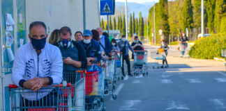 Eine lange Menschenschlange steht vor einem Supermarkt und wartet mit Einkaufswagen wohl auf Einlass. Die Schlange zieht sich bis über den Parkplatz und um eine Gebäudeseite herum.