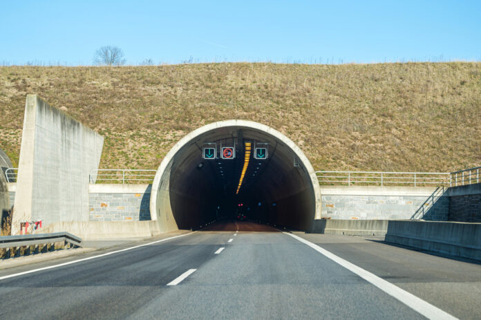 Einfahrt in einen dunklen Tunnel oder Autobahntunnel mit Beschilderung, Verkehrszeichen, Beleuchtung und Ampeln. Die Straße, die durch den Tunnel führt, ist zweispurig.