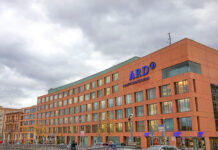 Das Gebäude des ARD-Hauptstadtstudios mit dem Logo der ARD in Blau am oberen Teil des Gebäudes. Das Gebäude aus Backstein ist riesig und beherbergt viele Räume. Vor dem Gebäude laufen einige Menschen.