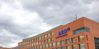 Das Gebäude des ARD-Hauptstadtstudios mit dem Logo der ARD in Blau am oberen Teil des Gebäudes. Das Gebäude aus Backstein ist riesig und beherbergt viele Räume. Vor dem Gebäude laufen einige Menschen.