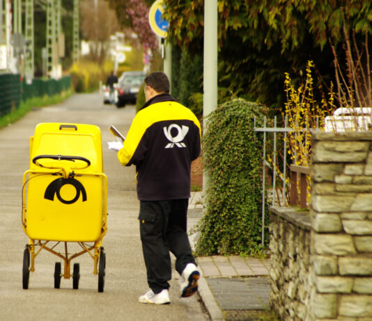 Ein Postbote der Deutschen Post steht neben seinem Postwagen, in dem sich viele Briefe befinden, auf einem Gehweg vor einem Haus und hält einige Briefe in der Hand. Er will sie wohl gerade an der richtigen Adresse in den Briefkasten werfen.