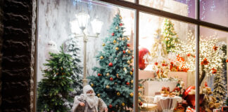 Es ist Weihnachten. Ein liebevoll dekoriertes Schaufenster in einem Kaufhaus zeigt eine Weihnachtsszene. Man sieht einen Weihnachtsmann und mehrere Weihnachtsbäume mit Weihnachtskugeln. Ein helles, warmes Licht zeigt einen mit Geschenken dekorierten Tisch.