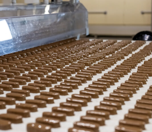 Schokoladen in Fabrik: Schokoladenriegel oder längliche Pralinen liegen auf einem Fließband und werden gerade mit Vollmilchschokolade überzogen. Danach gehen die Süßigkeiten in den Verkauf. ﻿