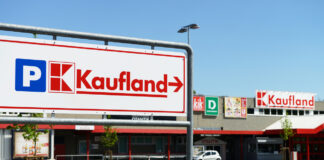 Ein großes Schild mit dem Logo von Kaufland. Dahinter liegt ein großer fast leerer Parkplatz. Wiederum dahinter ist die weitläufige Filiale des riesigen Supermarktes. Es sind zudem die Logos von Deichmann und kik zu erkennen.