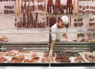 Eine gut bestückte Fleischtheke im Supermarkt. Ein Mitarbeiter nimmt die Waren aus der Kühltheke und im Hintergrund hängen allerhand Wurstwaren und Fleischerei-Produkte bereit. Die Umgebung ist steril, wie man es von einer Fleischerei erwartet.