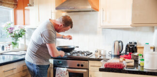 Ein Mann steht am Herd und kocht. Es steht eine Pfanne auf der Herdplatte und er streut wohl gerade ein Gewürz in das Essen. Am Griff des Backofen hängen zwei Ofenhandschuhe.