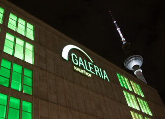 Das Galeria Kaufhof Hauptgebäude in Berlin. In der Dunkelheit leuchtet das Galeria-Logo in Grün und weiß. In den Fenstern des großen Gebäudes strahlt grünes Licht nach draußen.