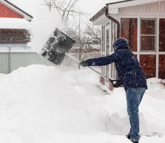 Ein Mann schippt Schnee, der einige Zentimeter hoch vor seinem Haus und auf dem Gehweg liegt. Auch auf dem Dach eines Hauses im Hintergrund liegt extrem viel Schnee.