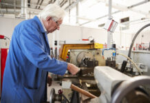 Ein alter Mann arbeitet in einer Halle oder Werkstatt an einer Maschine. Er führt etwas in die Maschine hinein. Der Mann trägt einen Blaumann und geht in seinem hohen Alter noch einer schweren körperlichen Arbeit nach.