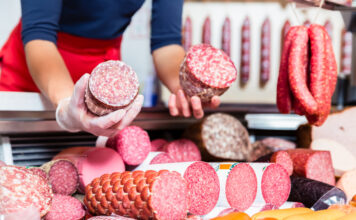 An der Wursttheke einer Metzgerei hält eine Frau zwei verschiedene Wurstsorten in die Kamera. Es handelt sich dabei um verschiedene Salami-Sorten. Die Wurst ist frisch aufgeschnitten und in der Auslage liegen noch viele andere Sorten von Fleisch.