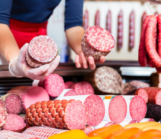 An der Wursttheke einer Metzgerei hält eine Frau zwei verschiedene Wurstsorten in die Kamera. Es handelt sich dabei um verschiedene Salami-Sorten. Die Wurst ist frisch aufgeschnitten und in der Auslage liegen noch viele andere Sorten von Fleisch.