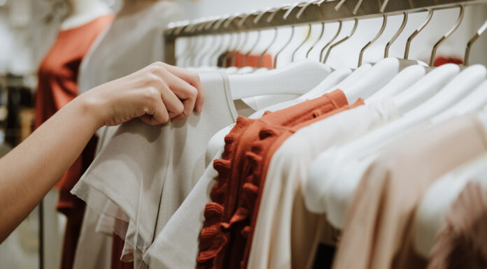 Eine Hand sucht an einer Kleiderstange Kleidung aus. Eine Person greift sich eines der Kleidungsstücke auf der Stange. Es handelt sich hierbei um neuwertige Mode.