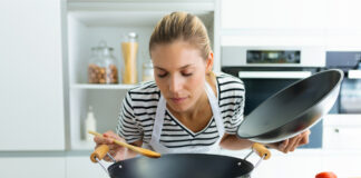 Eine Frau probiert das selbst gekochte Essen aus einem Wok, der auf einer eingeschalteten Herdplatte steht. In der linken Hand hält sie den Deckel. In der rechten nimmt sie etwas von dem Essen auf den Kochlöffel.