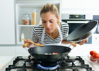 Eine Frau probiert das selbst gekochte Essen aus einem Wok, der auf einer eingeschalteten Herdplatte steht. In der linken Hand hält sie den Deckel. In der rechten nimmt sie etwas von dem Essen auf den Kochlöffel.