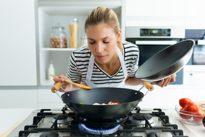 Eine Frau probiert das selbst gekochte Essen aus einem Wok, der auf einer eingeschalteten Herdplatte steht. In der linken Hand hält sie den Deckel. In der rechten nimmt sie etwas von dem Essen auf den Kochlöffel.