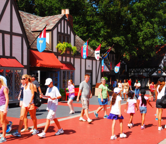 Zahlreiche Gäste laufen im Sommer und in entsprechend sommerlicher Kleidung in einem Freizeitpark umher. Im Hintergrund liegen Häuser im Fachhaus-Stil. Es wehen Fahnen im Wind und orange Markisen sind herabgelassen.