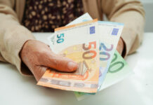Eine Seniorin sitzt am Tisch und hält Geldscheine in der Hand und trägt dabei eine Strickjacke und ein geblümtes Oberteil.
