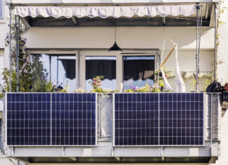 Auf einem Balkon befindet sich ein Balkonkraftwerk mit vielen Solarzellen, die praktisch einen Sichtschutz um den Balkon herum bilden. Dahinter sieht man ein paar Bäume, eine Markise und eine Lampe, die von der Decke herunterhängt.