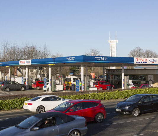 Blick auf eine Tankstelle. Im Vordergrund sieht man eine Straße mit vielen Autos, die daran vorbeifahren. Die Ladesäulen an der Tankstelle selbst sind ebenfalls voll gestellt mit Autos.