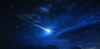 Ein Komet fliegt am dunkelblauen Himmel in der Nacht und leuchtet hellblau bis weiß auf. Vereinzelte Wolken lassen die Atmosphäre besonders mysteriös erscheinen.