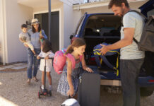 Eine Familie mit drei Kindern packt den Kofferraum ihres Autos. Der Vater lädt einen Kinderrucksack ein und direkt vor dem Auto stehen weitere Koffer und Gepäckstücke. Es scheint so, dass die Familie in den Urlaub fährt.