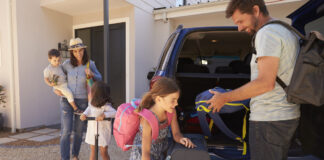 Eine Familie mit drei Kindern packt den Kofferraum ihres Autos. Der Vater lädt einen Kinderrucksack ein und direkt vor dem Auto stehen weitere Koffer und Gepäckstücke. Es scheint so, dass die Familie in den Urlaub fährt.