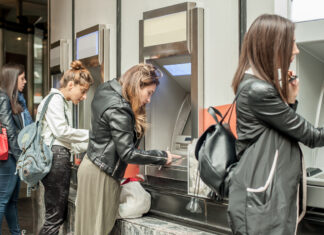 Frauen an einem Geldautomaten in einer Bank. Sie stehen jede an einem eigenen Automaten, tippen ihren Pin ein oder warten auf die Geldbewegung oder Auszahlung. Die Filiale ist hell beleuchtet.