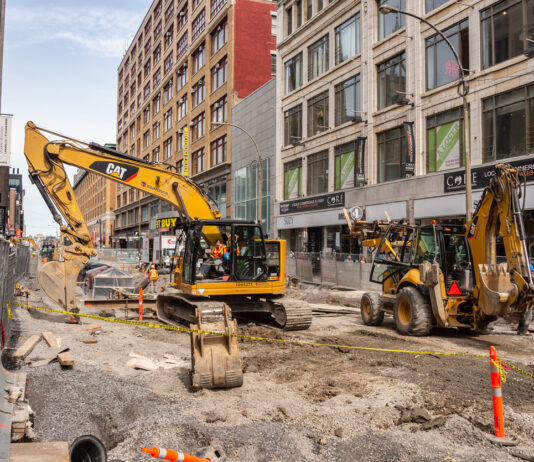 Ein Bagger auf einer Baustelle in Karlsruhe. Die Baumaschinen haben den gesamten Boden aufgerissen. Im Hintergrund Hochhäuser und große Gebäude. Die Baustelle ist abgesperrt.