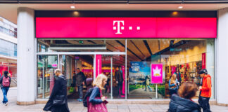 Eine Telekom-Filiale in einer Innenstadt. Der Laden des Handy- und Telefonanbieters ist in einem klassisch pinken Telekom-Farbton gehalten. Vor dem Geschäft laufen mehrere Passanten vorbei.