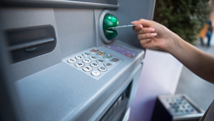 Eine Bankkarte wird in den Schlitz des Automaten gesteckt. Dieser ist mit Grün gekennzeichnet und soll so vor Datendiebstahl schützen. Der Fokus liegt klar auf dem Tastenfeld und dem Kartenschlitz des Automaten.