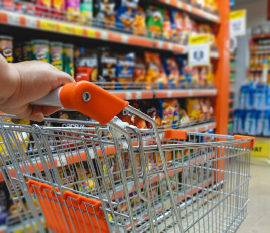 Ein leerer Einkaufswagen mit orangen Details wird von einer Person im Supermarkt geschoben. In den Regalen dahinter stehen verschiedene Lebensmittel in bunten Verpackungen.