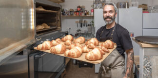 Ein Bäcker steht mit einem Backblech in der Backstube. Auf dem Backblech befinden sich verschiedene Backwaren wie Brötchen und Brot. Diese werden in der Bäckerei von der Bäckereifachkraft an die Kunden verkauft.