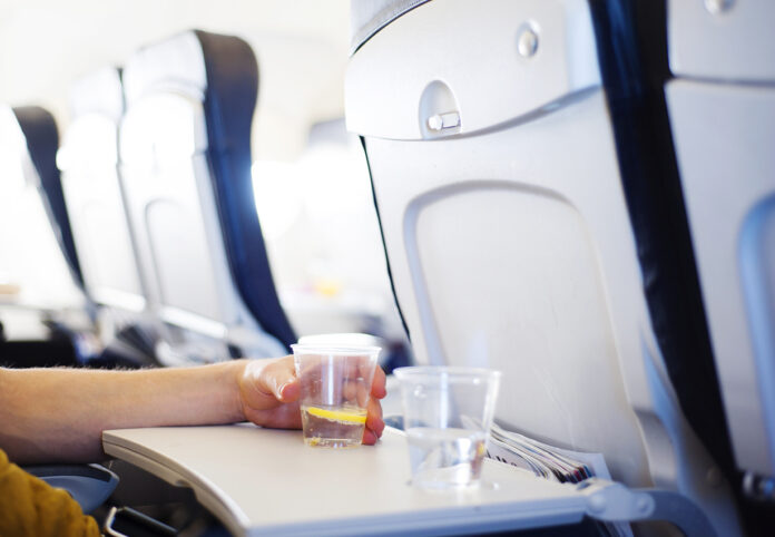 Ein Glas Wasser steht auf einem Tisch in einem Flugzeug. Ein Passagier oder Fluggast greift nach diesem Wasserglas, um zu trinken auf dem Flug.