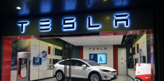 Ein weißes großes E-Auto von Tesla steht in einem Showroom hinter einem Schaufenster. Über dem Ausstellungsfenster prangt das Logo von Tesla. Das Fahrzeug ist ein E-SUV.