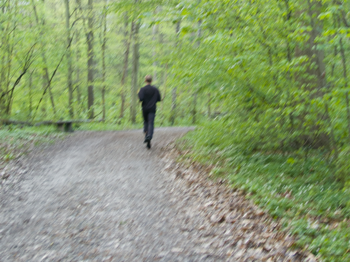 Mann läuft im Wald auf einem Waldweg. Am Rand stehen Bäume, weshalb es sich um einen Mischwald handelt. Es liegt Laub am Wegrand.