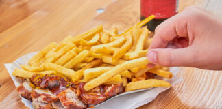 Hand greift zu Pommes mit Currywurst in einer Pappschale mit einer Cola daneben.